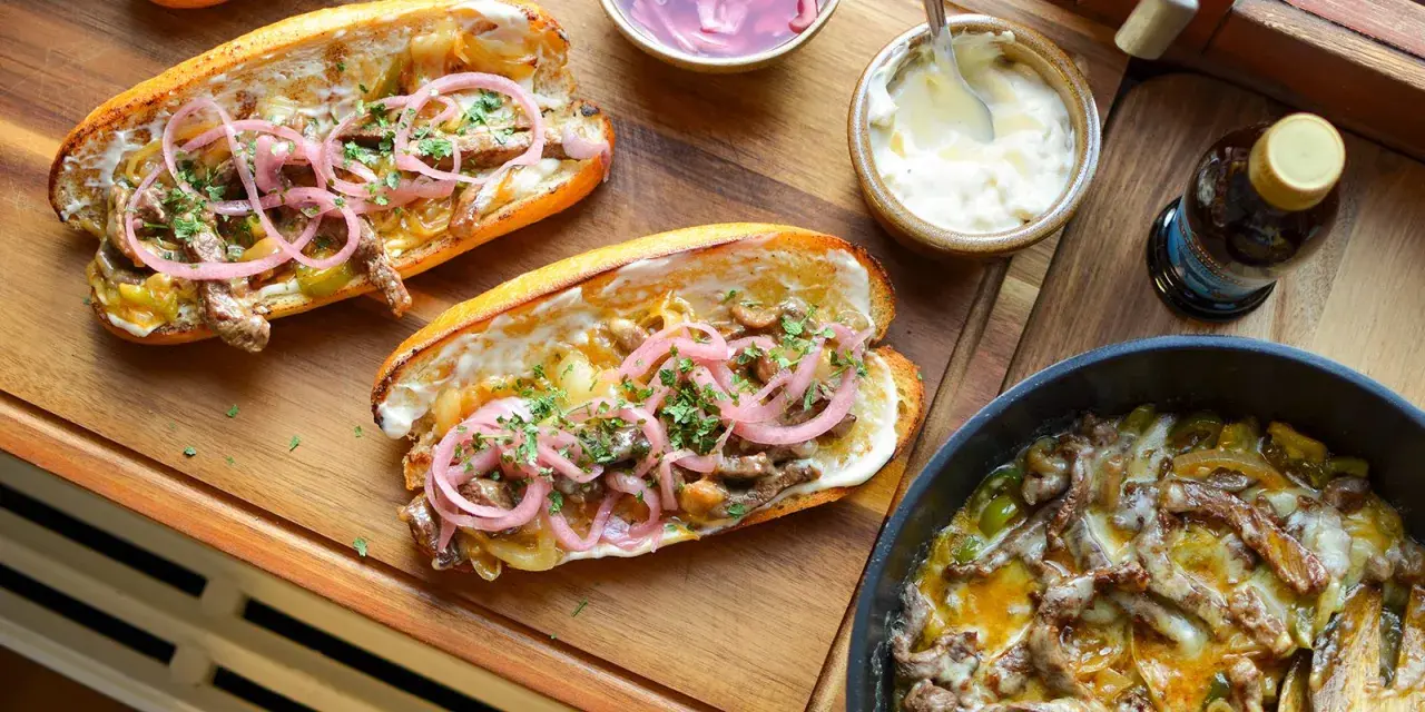 Två philly cheese steak-mackor med möra strimlor av kött, smält ost, lök och paprika, toppade med picklad rödlök och persilja.