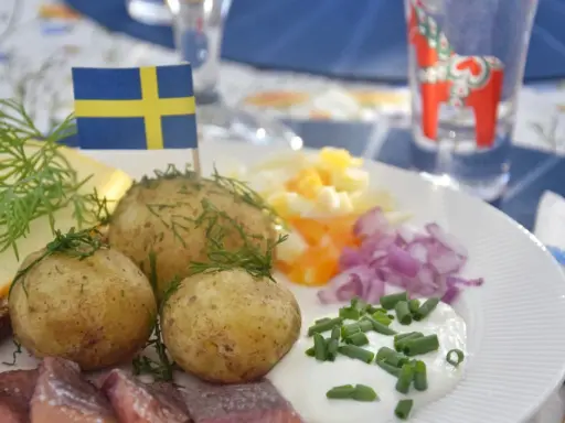 En tallrik med midsommar mat: sill, färskpotatis, ost, ägg och rödlök. En svensk flagga vajar stolt.