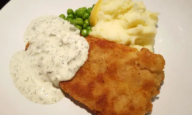 Panerad fisk med krämig kall sås till, serverad med potatismos, gröna ärtor och en citronklyfta.
