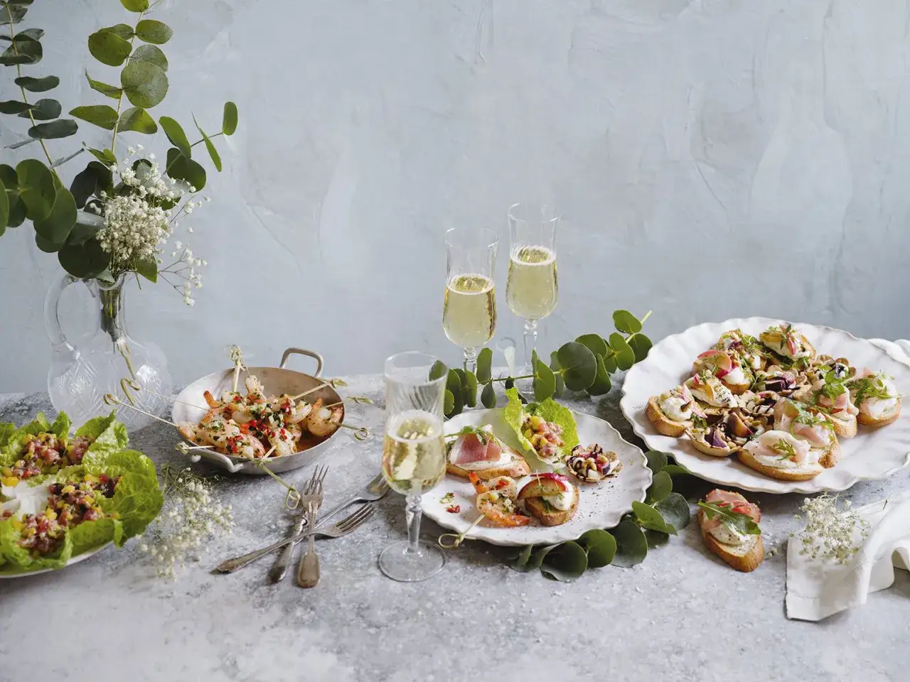 Festliga tilltugg till bubbel: r&auml;kor p&aring; spett, bruschetta med olika p&aring;l&auml;gg och sallad i salladsblad. Tv&aring; glas bubbel st&aring;r redo.