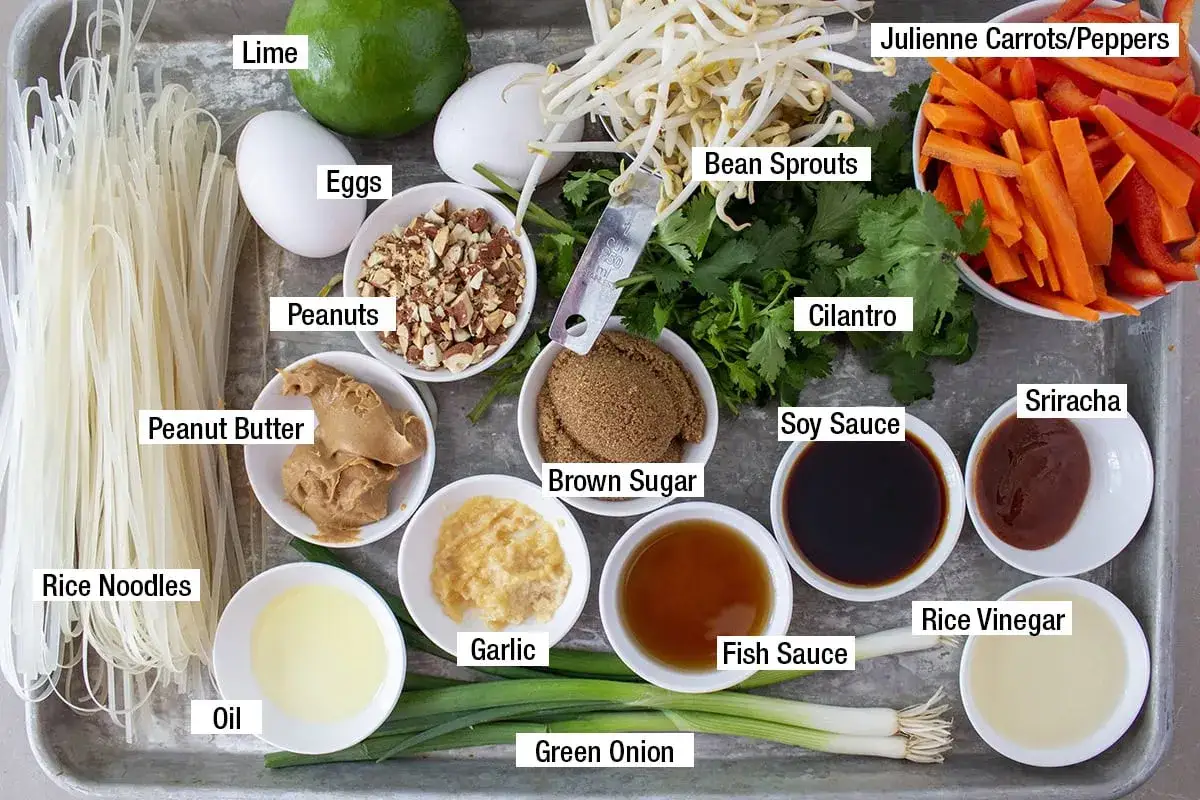 Ingredienser för en smakrik pad thai sås: risnudlar, jordnötssmör, lime, ägg, bönskott, koriander, sriracha, sojasås, fisksås, risvinäger, vitlök, socker, olja, jordnötter, morötter, paprika och salladslök.