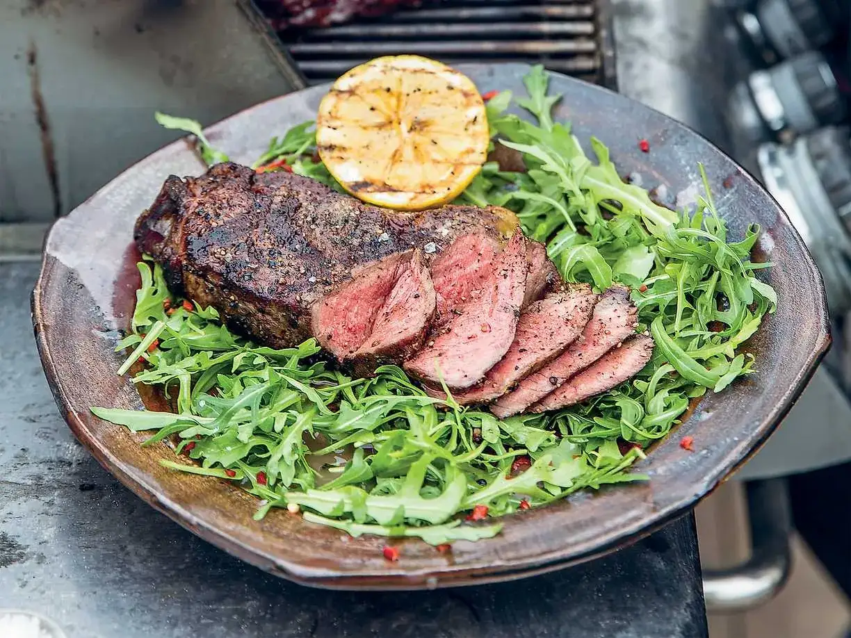 Perfekt grillad ryggbiff, skivad och serverad p&aring; en b&auml;dd av ruccola med en grillad citronklyfta.