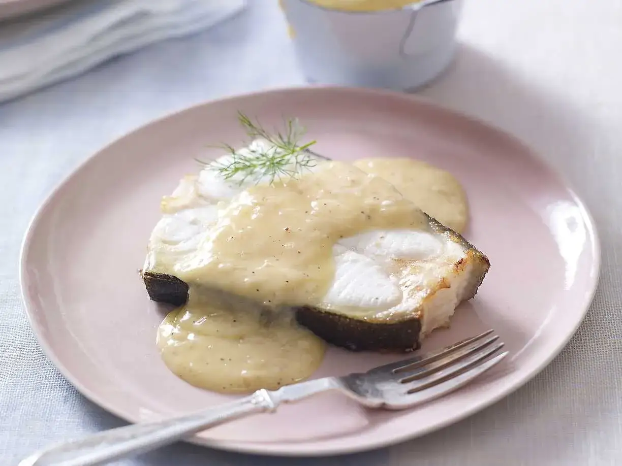 En bit stekt fisk med en kr&auml;mig beurre blanc-s&aring;s och dill p&aring; en rosa tallrik.