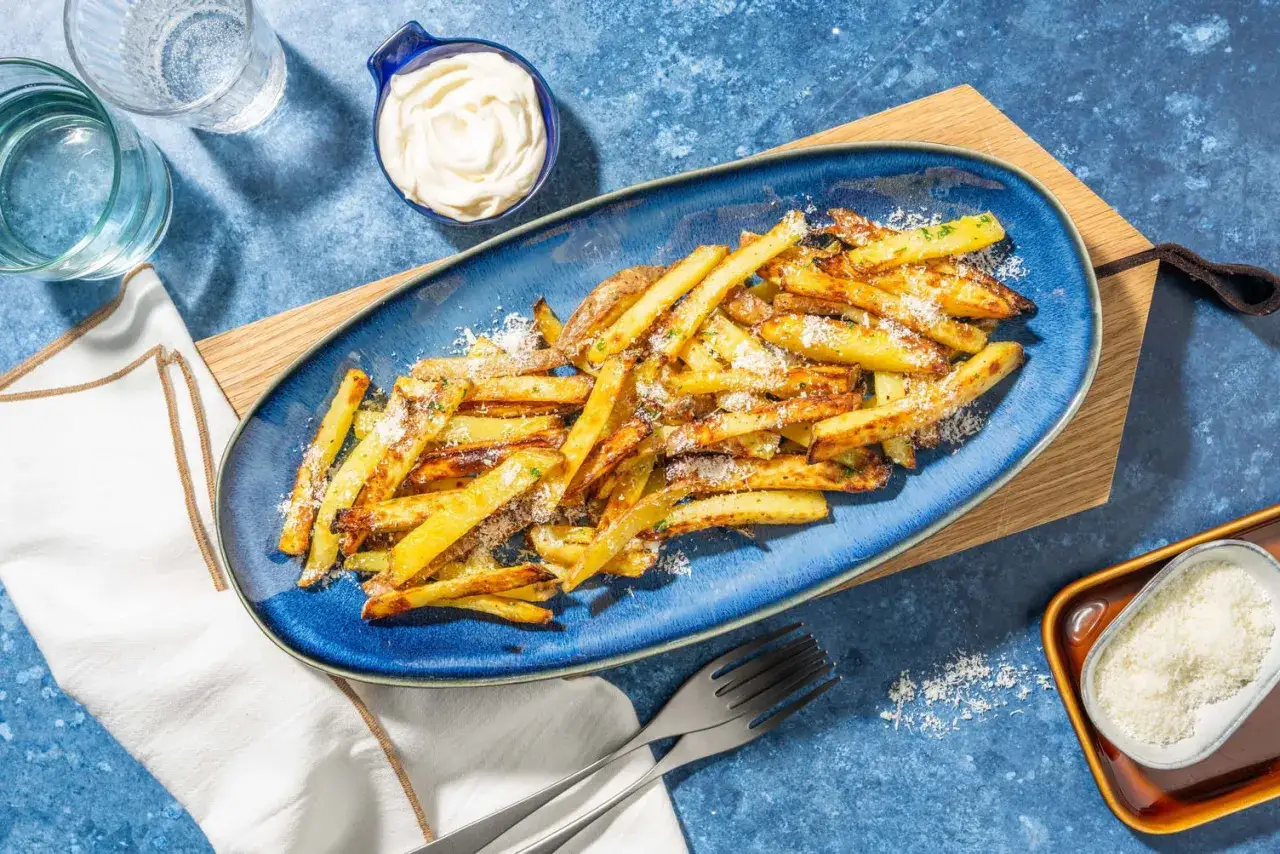 En bl&aring; tallrik med gyllene pommes frites, toppade med riven ost och &ouml;rter. Bredvid finns en sk&aring;l med majonn&auml;s och ett glas vatten.