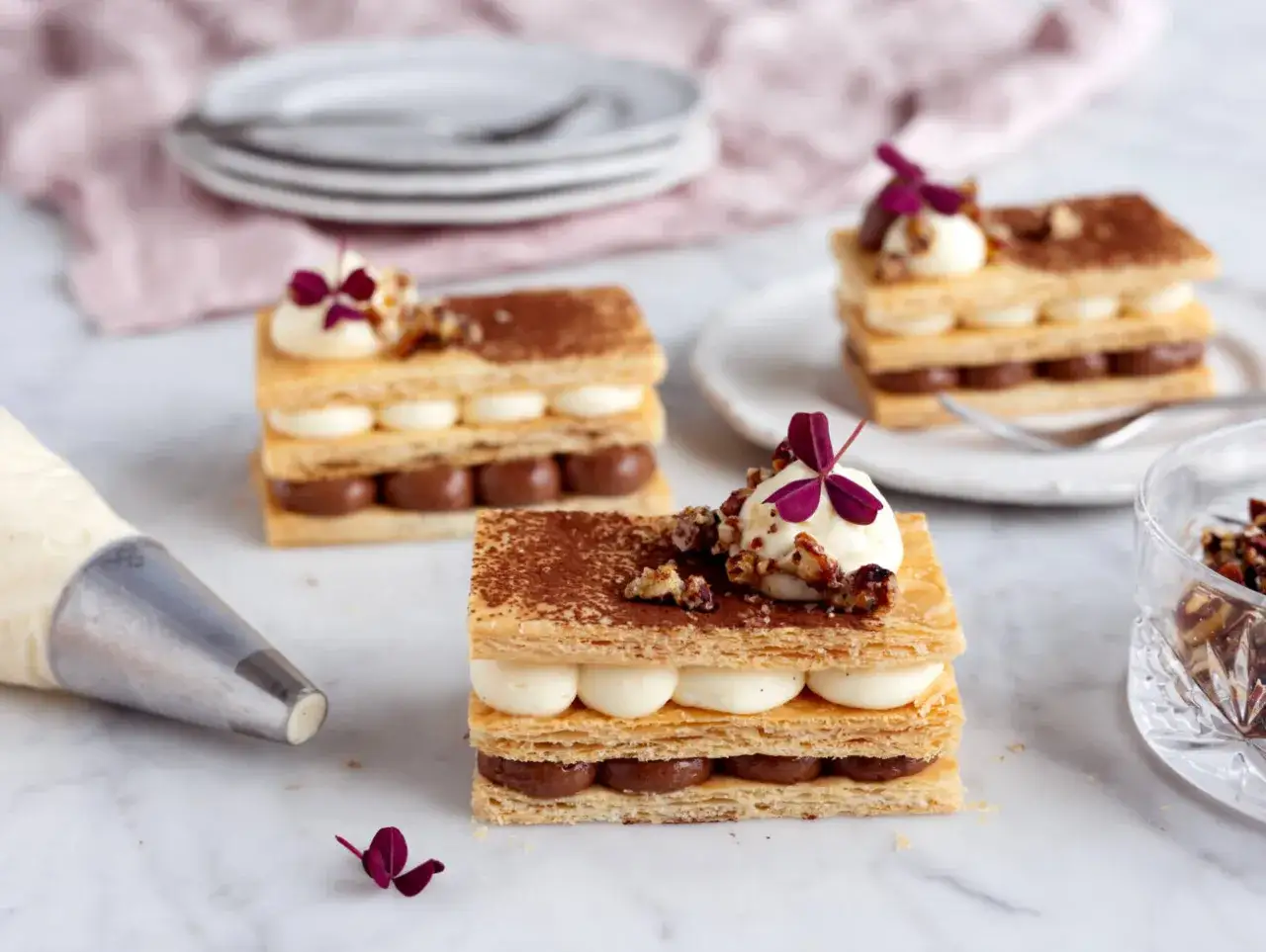 Tre eleganta millefeuille-desserter med lager av krispigt bakverk, chokladkr&auml;m och vaniljkr&auml;m, toppade med n&ouml;tter och en lila blomma.