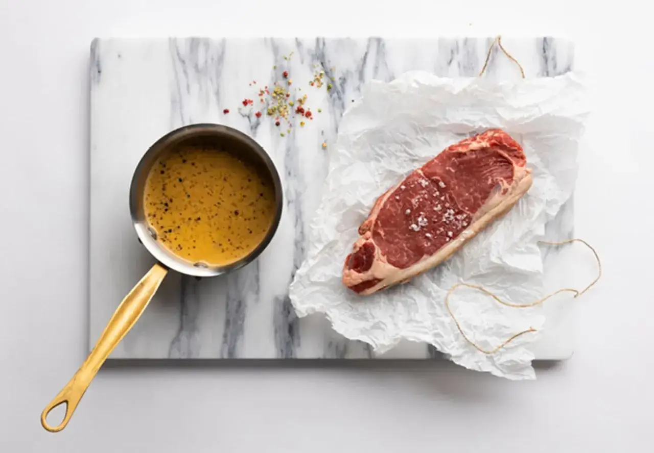 En rå biff med salt och peppar ligger på smörpapper bredvid en kastrull med en gyllene pepparsås. Recept på pepparsås här!